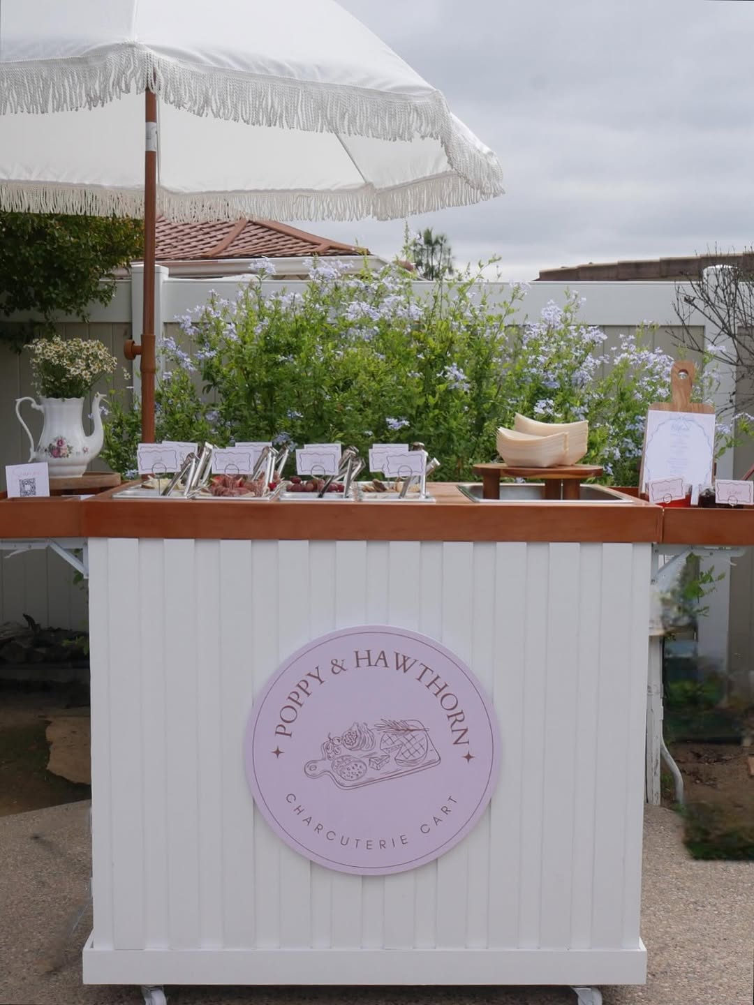 The Poppy & Hawthorn charcuterie cart set up outdoors with parasol and florals